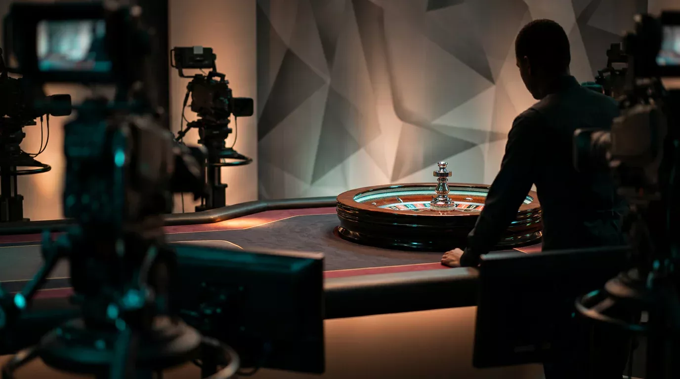 Estudio de casino en vivo con mesa de ruleta europea, cámaras de televisión y silueta de un crupier al fondo